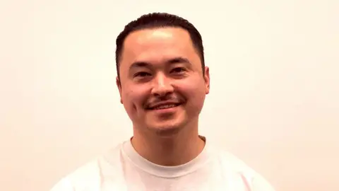 Konrad Cheng is wearing a plain white short-sleeved t-shirt. He has black hair, brought behind him. He is standing in front of a white wall and is centred in the frame, from the shoulders upwards.