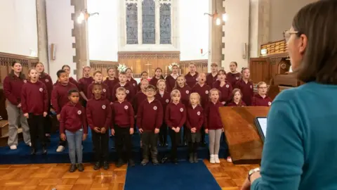 Children wearing purple jumpers sing in a church, led by a woman stood at a lectern.