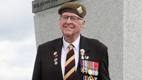 Richard Brock smiling in a white shirt and black suit jacket with his regimental cap, tie and medals.