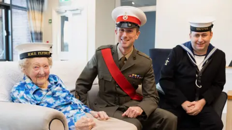 St Monica Trust Margaret Grey wearing a blue and white patterned blouse and a white and black naval cap with yellow embroidered letters. She is sitting on a cream sofa, looking at the camera. Beside her is John Morrish, wearing a khaki naval uniform with a red sash, black, red and white cap, and colourful naval badges on his right breast pocket. On the right is another naval officer, wearing a black uniform and a white cap. 