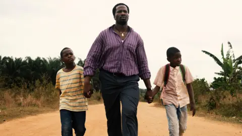 Actor Sope Dirisu walks down a dusty road whole holding the hands of young actors Godwin Chiemerie Egbo and Chibuike Marvellous Egbo. Sope is wearing a purple striped shirt and jeans. The boys are wearing jeans and T-shirts. 