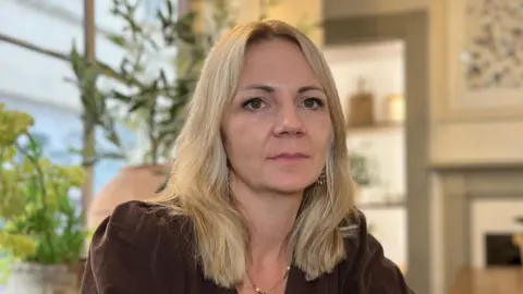 Chloe has shoulder-length light hair seated at a wooden table in a softly lit dining room, with plants, a window, and framed artwork in the background.