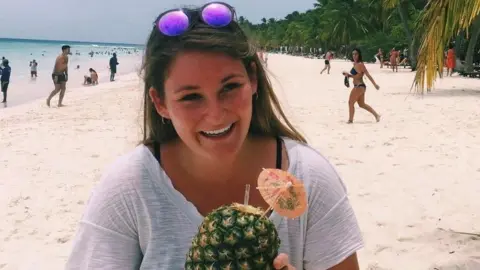 Family handout Emily Oliver on a sandy beach, smiling and holding a pineapple