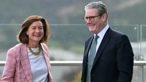 Eluned Morgan and Sir Keir Starmer stood next to each other with a steel bar behind them. Morgan is wearing a pink jacket and white top while Sir Keir is wearing a black suit, white shirt and green tie.