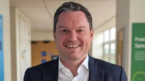 Gloucestershire Hospitals NHS Foundation Trust A picture of Kevin McNamara in a corridor, he is wearing a navy blazer and a white shirt, and smiling widely.