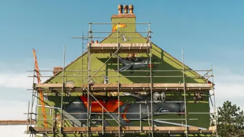 Greyhound Scaffolding around the side of a green coloured pub, with the painting of a startled looking greyhound behind.