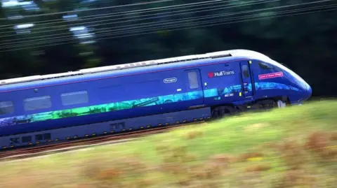A Hull Trains train in dark blue named Land of Green Ginger with images of the city on a strip down the side speeding along a railway line with the grass in front of it and the overhead wires blurred