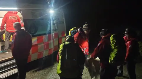 Dartmoor Search and Rescue Team Plymouth Members of Dartmoor Search and Rescue Team Plymouth gather outside of the rescue van on Tuesday evening. The team are wearing warm hats and coats. It is dark. One team member is inside the van looking at a computer screen which appears to show a map. Other members of the team are gathered around a paper map. A head torch is being used to illuminate the map.