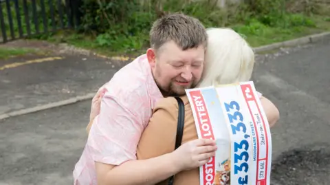 People's Postcode Lottery A man and a woman are hugging whilst standing in a street. The woman is wearing a brown coat and the man is wearing a pink shirt and holding a piece of paper which has been styled to look like a cheque.