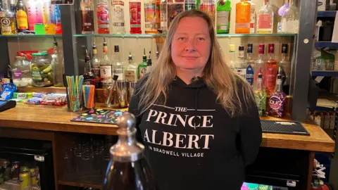 Amy Holmes/BBC A woman standing behind a bar with another wooden bar top behind her along with various bottles of spirits, snacks, straws etc.