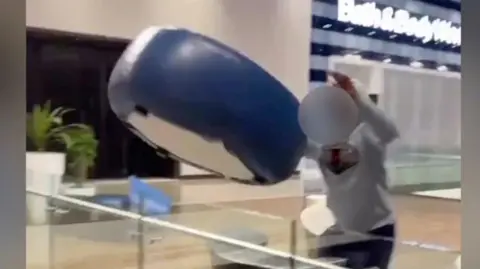 A teenager wearing a grey hoodie and black trousers, is standing over a glass balustrade, having thrown a large blue seat, which is in mid-air. His face is blurred.