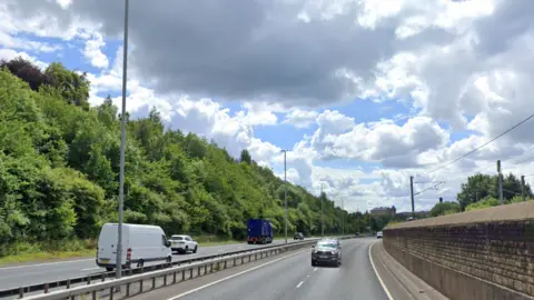 Google A white van, three cars and a blue truck drive along the A650. There is a railway line to the right and a line of trees and bushes to the left.