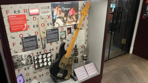 BBC A black and gold bass guitar in a museum display box. Brit and Ivor Novello awards are also visible.