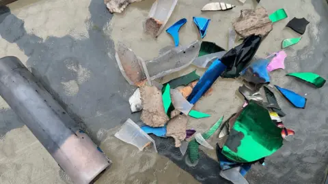 BBC/Leanne Brown Remains of fireworks, including a plastic tube and pieces of brightly coloured, sharp-edged plastic, displayed on a garden table. The colours include blue, pink and green.