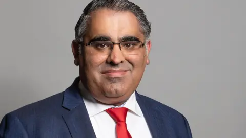 Head and shoulders shot of MP Tahir Ali. He is wearing half-rimmed glasses, a blue suit and a red tie