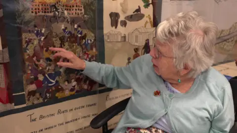 Dorothy Nicholls wearing a pale blue cardigan with a rose brooch. She has short white hair and glasses, and is sitting in a black wheelchair beside the framed tapestry. She has her right forefinger extended, pointed at the detailed stitching of a large picture, depicting the Queen Square Riots of 1831.