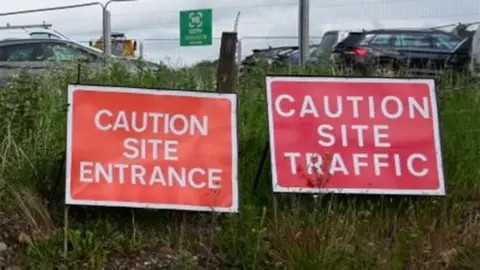 Two rectangular signs, with white text on red, by a grass verge. The one on the left says "caution site entrance" and the one on the right says "caution site traffic"