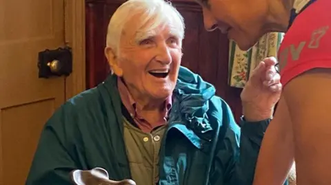 BBC Gordon Green, with medium length white hair, wearing a green waterproof coat, smiling at BBC newsreader Sophie Raworth, who had taken part in the man v horse race