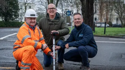Gloucestershire County Council From left to right, Alan Williams, Kaarbon Tech, Councillor Julian Tooke, Cabinet Member for Business, Economic Development and Matt Smith, Programme Manager for River Severn Partnership - Advanced Wireless Innovation Region, pose with one of the sound wave detectors on the side of a road.