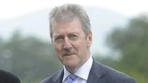 Mr Travers has short grey hair and a moustache and is wearing a grey suit with a blue striped tie and a white shirt.