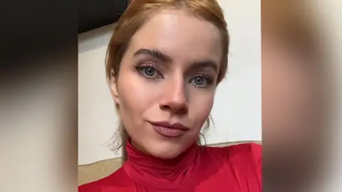 Close up of Ana Machado with long light brown hair wearing a red polo neck top sitting on a beige sofa. She is smiling. 