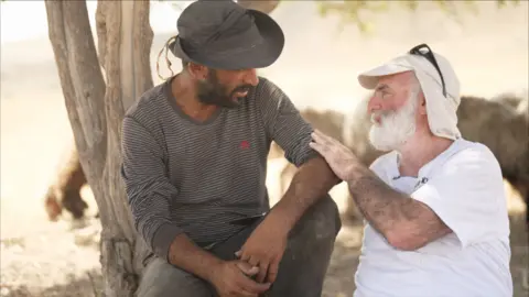 Ahmad, a Palestinian farmer wearing a long-sleeved T-shirt and a hat, sits with Gil and a white-bearded man. Gil has his hand on Ahmed's arm and they sit under a tree talking.