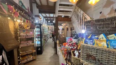 Shop floor with wooden shelves and ceiling beams, stone walls, bunting, various goods on display