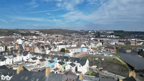 An aerial photo of Aberystwyth. Rows of terraced houses can be seen leading to the river bank, with a bridge going over the river in the horizon. There are four boats moored up by the river bank. 