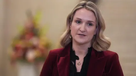 Helen McEntee is looking away from the camera. She has shoulder length blonde hair. She is wearing a red jacket and black blouse.