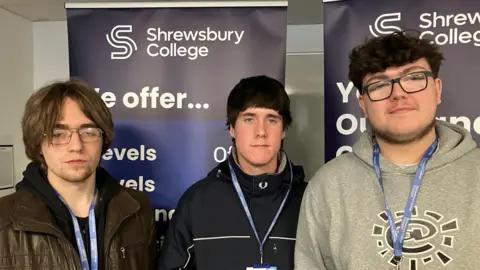 Three young people with short dark hair one has a brown leather jacket and glasses, another has a black top and the third has a grey top and glasses