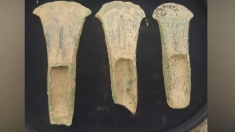 Peter Major Three axe heads recovered by Peter Major. They are of a pale yellow colour with some green parts. They are laid on a black surface. 