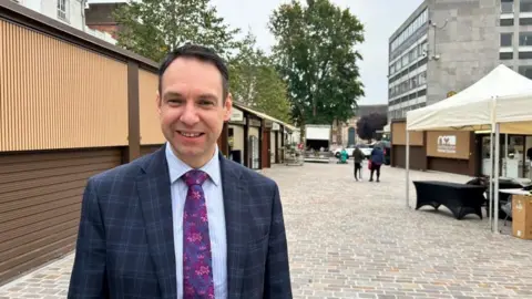 Tom Toseland/BBC Dan Lister with short dark hair, wearing a blue jacket and purple tie and smiling with the new brown market stalls in the background