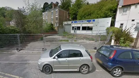 Two cars parked by a fenced-off area of concrete. A building is behind the former forecourt with "Express Shop" written on it.