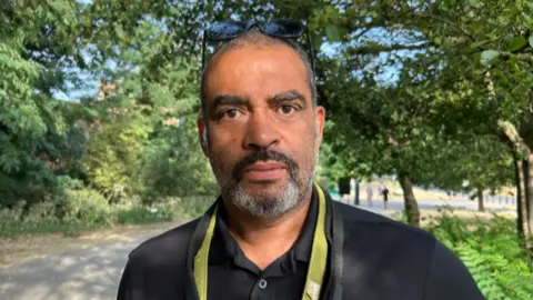 A person wearing a black polo shirt stands on a tree-lined path in a park. A pair of sunglasses rests on the person's head and a green lanyard hangs around the neck.