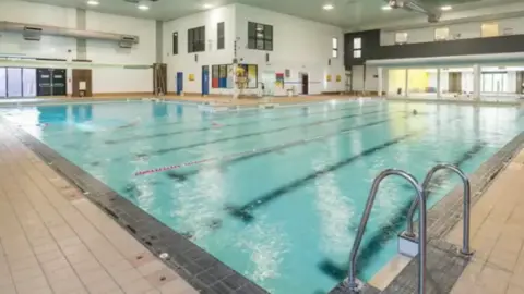 A leisure centre swimming pool. The white room has a large swimming pool which has dark blue lanes and water which looks light blue. The area around the pool is covered in cream coloured tiles and a there is a metal ladder in the front right corner leading into the water.