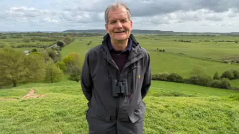 A man wearing a grey coat over a purple jumper, with binoculars around his neck. He's smiling at the camera, backed by green fields and a grey sky.