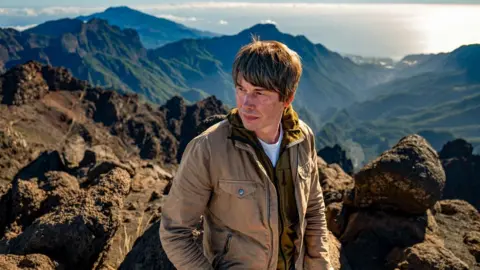 Prof Brian Cox sits on a hillside. He is wearing a brown jacket and jeans and has a pensive look on his face. The Sun is shining brightly in the background.