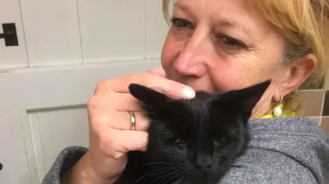 Clay Cottage Cattery A blonde woman wearing a grey hooded jacket holds a black cat in front of a white door