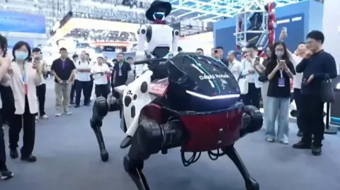 A humanoid robot wearing a cowboy hat sits on a robot horse as human spectators watch on