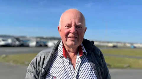 Chris Le Tissier standing outside looking at the camera. He is wearing a grey coat on a white striped shirt. He is mostly bald with short white hair.