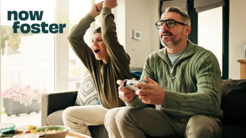 A promotional image for Now Foster. A boy and a man sit together on a settee playing video games. The man, who is dressed in a jumper and wears glasses, is holding a controller and concentrating. The boy, dressed in an olive-coloured long-sleeved top, has his arms in the air in celebration.