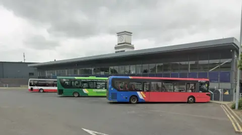 Three buses are parked outside Stanley bus station. It is a grey glass building with a white clock tower rising above it. 