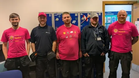 Brain Tumour Research Liam Harvey wearing a pink Brain Tumour Research T-shirt. He is standing in a line alongside four of his male colleagues from work. They are all wearing black cargo trousers and are standing in an office room in front of blue metal lockers.