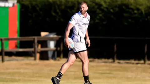 LooseHeadz Former rugby player George Kruis - a man with short brown hair and beard, and wearing a white kit - turns to his right and laughs as he holds a rugby ball by his side while walking on the pitch.