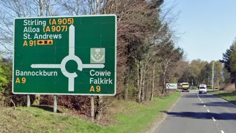 Google Roundabout sign on the A91. there are a couple of vehicles on the road and trees on either side of the road 