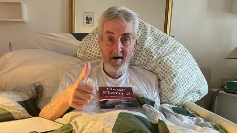 A man with short grey hair and a beard, is wearing a white T-shirt. He is sitting up in a bed, with a pillow behind him, and is looking at the camera and giving a thumb's up with his right hand. On his chest is a book called "Dear Flora 2".