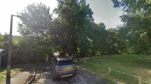 A screengrab taken from Google images which shows a footbridge in the distance. There is a silver car parked next to a patch of grass.