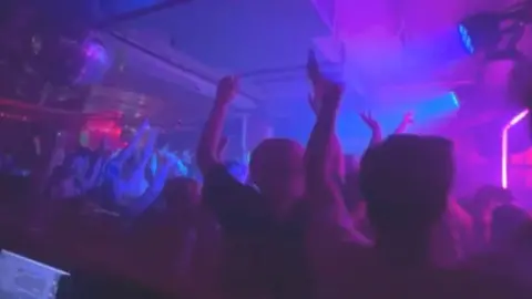 Kate Hodgkinson Dancers at a Cobalt Studios concert are waving their hands in the air. The venue is lit by blue and purple lights