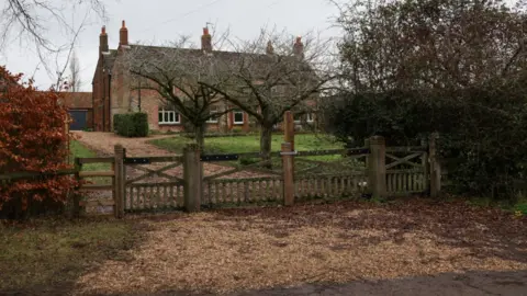 PA Media A view of red brick farmhouse, pictured from a road. It is behind trees and a wooden gate
