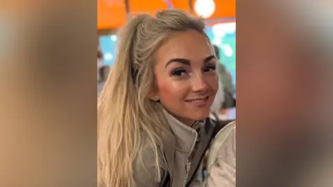 Francesca Moriarty Francesca Moriarty is in the middle. She has long blonde hair in a ponytail and is wearing a beige jacket and beige jumper. 